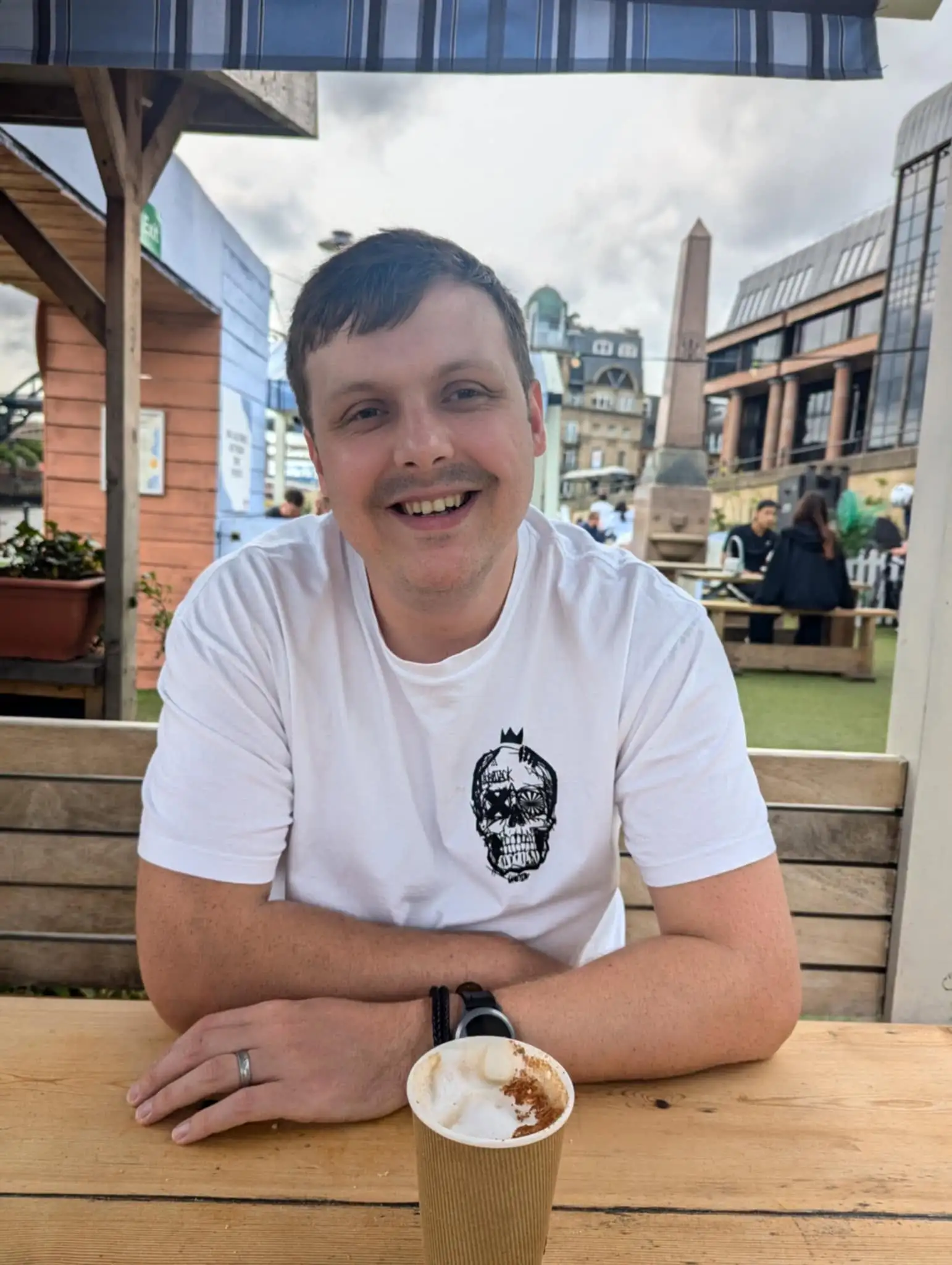 Matt Andrews-Mills in a white t shirt, sat at a table with a coffee